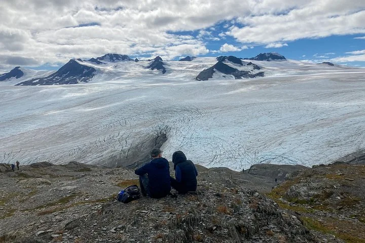Due persone sedute su una sporgenza rocciosa che domina un enorme ghiacciaio.