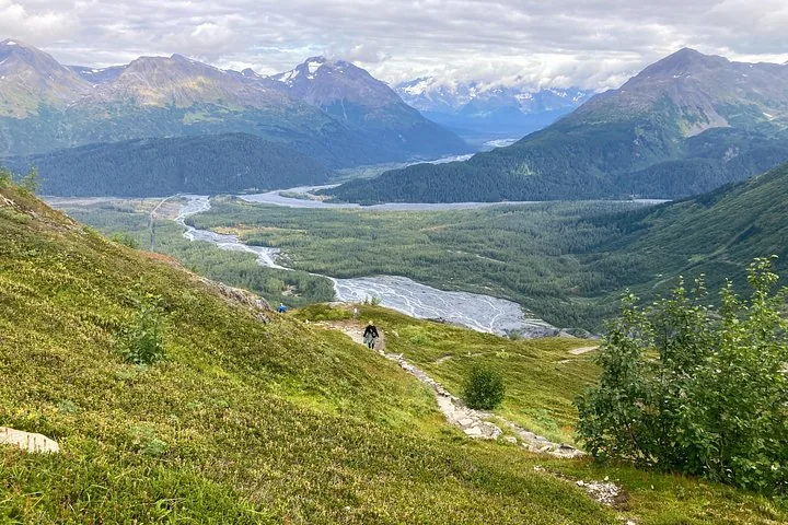 Un escursionista in lontananza che cammina su un sentiero che domina una valle verde e un fiume.